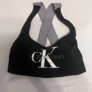 Svart linnetopp från Calvin Klein - Svart linnetopp från Calvin Klein med stort vitt CK-tryck framtill. Toppen har breda grå axelband som korsas i ryggen för en sportig look. Knappast använd 