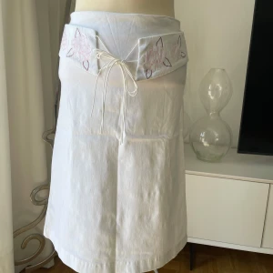 Vit kjol från Vero Moda - Supersöt vit kjol från Vero Moda med broderade blommor i rosa och vinrött på linningen. Kjolen har knytband framtill och är tillverkad i ett mjukt bomullstyg. Perfekt för en fräsch och somrig look! Midjemått 74 cm