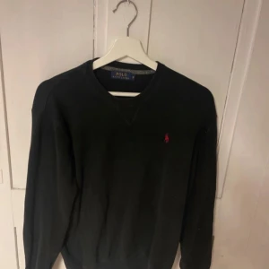 Mörk tröja från Polo Ralph Lauren - Klassisk mörk tröja från Polo Ralph Lauren med rund halsringning och röd broderad logga på bröstet. Tröjan har långa ärmar och ribbade muddar vid ärmslut och nederkant. Perfekt för en stilren och enkel look.