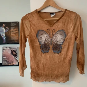 Brun meshtröja med fjäril - Superfin brun meshtröja köpt i en vintagebutik i Mexiko med stort fjärilstryck i blått och grått framtill. Tröjan har lång ärm, vågig kant nertill och vid ärmslut samt en liten v-ringning vid halsen. Tunn och lätt genomskinlig