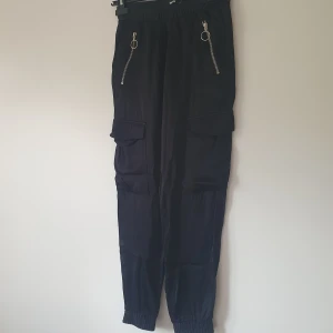 Svarta cargopants från Bik Bok XS - Svarta cargopants från Bik Bok i storlek XS. Byxorna har resår i midjan och vid bensluten, två stora fickor på sidorna och dragkedjor med ringdetaljer framtill. Perfekta för en chill och trendig look.  Helt skick dock små noppror