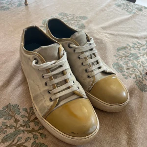 Lanvin sneakers med beige och gulddetaljer - Snygga sneakers från Lanvin i beige canvas med glansiga guldfärgade tår och detaljer. Klassisk rund tå och robust sula. Perfekta för dig som gillar en stilren men ändå unik look. säljs utan box i storlek 42 men passar till 41 och 42,5. Frågor? Skriv