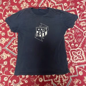  GANT t-shirt  - Blå t-shirt från GANT med stort vitt tryck på bröstet i form av ett sköldmärke och texten GANT. Klassisk rund hals och korta ärmar. Skön och enkel modell som passar till det mesta. Materialet känns mjukt och troligtvis i bomull.