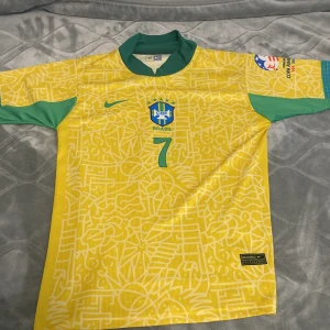 Brasilien Nike fotbollströja gul/grön M - Snygg Brasilien fotbollströja från Nike i storlek M. Tröjan är gul med gröna detaljer, har ett abstrakt mönster och nummer 7 på både fram- och baksidan samt namnet Juliano. Klassisk krage och korta ärmar. Perfekt för dig som älskar fotboll och vill sticka ut.