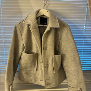 Ljusgrå ulljacka från 157, XS - Snygg ljusgrå jacka från 157 i ullblandning med klassisk krage, stora fickor framtill och knäppning med tre knappar. Jackan har en rak och lite boxig passform som ger en modern vibe. Perfekt för dig som gillar stilrena och enkla plagg.