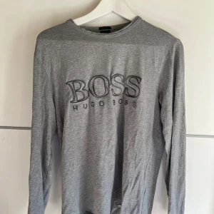 Grå långärmad tröja från Hugo Boss - Snygg grå långärmad tröja från Hugo Boss med stort broderat BOSS-tryck på bröstet. Tillverkad i mjuk bomull, perfekt för en clean och stilren look. Klassisk rund halsringning och relaxed passform.