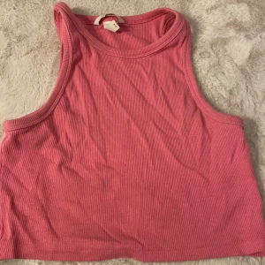Rosa ribbad croppad linnetopp H&M - Säljer en rosa croppad linnetopp från H&M. Toppen är ribbad och ärmlös med breda axelband och rund halsringning. Perfekt för varma dagar eller att styla lager på lager. Enkel och snygg att matcha med jeans eller kjol.