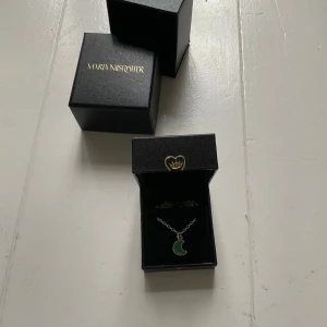 Grönt månhalsband från Maria Nilsdotter - Unikt halsband från Maria Nilsdotter med en silverkedja och ett grönt månhänge. Kedjan har en liten bricka med texten 'DOTTER'. Perfekt statement-piece för dig som gillar coola detaljer och snygga smycken.