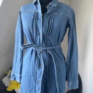 Jeansklänning XOXO Friends med bälte - Snygg jeansklänning från XOXO Friends i klassisk blå denim. Klänningen har långa ärmar, knappar framtill och ett matchande bälte i midjan. Snygga sömdetaljer framtill ger extra stilpoäng. Perfekt för dig som gillar en chill men trendig look.