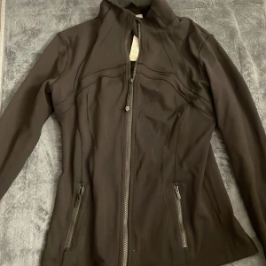 Svart vindjacka från Lululemon, strl 10/XL - Snygg svart vindjacka från Lululemon define med figurnära passform och hög krage. Jackan har dragkedja framtill, två fickor med dragkedja och långa ärmar. Perfekt för träning eller när du vill ha en sportig look.
