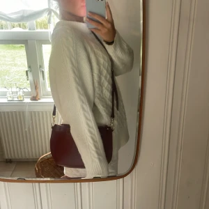 Mörkröd axelväska i skinn - Säljer en stilren mörkröd axelväska i slätt skinn med guldfärgade detaljer och dragkedja upptill. Väskan har en enkel, rundad form och justerbar axelrem. Perfekt storlek för det viktigaste och passar till många olika outfits.