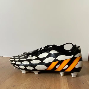 Adidas Predito 14 fotbollsskor svart/vit - Snygga Adidas Predito 14 fotbollsskor med svart bas, vita geometriska mönster och orangea ränder på sidorna. Skorna har snörning, platt sula med fasta dobbar och är tillverkade i syntetmaterial. Perfekta för dig som vill sticka ut på planen. Inhandlades för många år sedan och bara använts ett fåtal gånger. Ganska sällsynta och väldigt coola :)