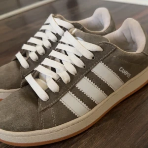Adidas Campus sneakers i grå mocka - Snygga Adidas Campus sneakers i grå mocka med klassiska vita ränder och vit sula. Skorna har rund tå, platt sula och vita snören. Perfekta för dig som gillar en clean och tidlös stil med streetkänsla.