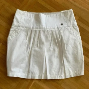 Vit kortkjol från Diesel med knappar - Vit kortkjol från Diesel i bomull med snygga detaljer som knappar bak, fickor och veck framtill. Kjolen har bred midja och coola sömmar som ger en fräsch och modern look. Perfekt för dig som gillar stilrena och trendiga plagg.