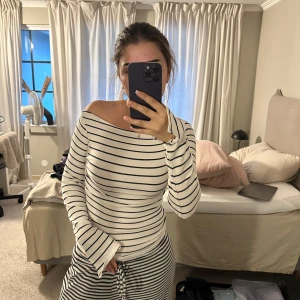 Randig långärmad tröja i bomull - Vit långärmad tröja med svarta horisontella ränder och offshoulder-skärning. Tröjan är figurnära och har långa ärmar med lätt utsvängda muddar. Tillverkad i mjuk bomull, perfekt för en chill och stilren look.