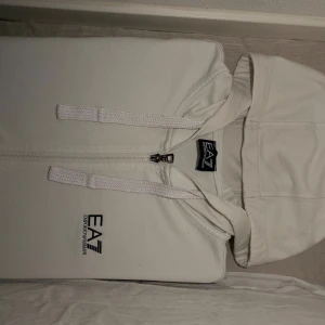 Vit hoodie från EA7 Emporio Armani - Säljer en stilren vit hoodie från EA7 Emporio Armani med dragkedja och huva. Tröjan har snörning vid huvan och svart broderad logga på bröstet. Perfekt för dig som gillar exklusiva streetwear-vibbar och vill ha något bekvämt och snyggt.