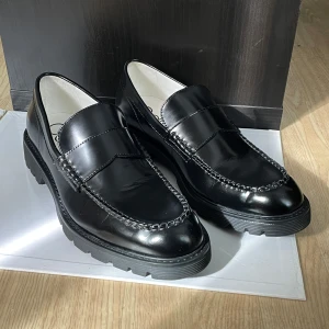 Svarta loafers från Calvin Klein - Chunky svarta loafers från Calvin Klein i glansigt skinn med markerade sömmar och kraftig platt sula. Klassisk penny loafer-design med rund tå och stilren look. Perfekta för dig som vill ha en clean och modern vibe. Endast använda en gång, så skicket är mycket bra. Nypris 2000kr. 