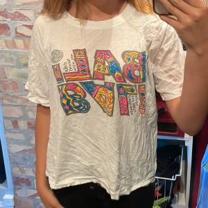 Vit t-shirt med färgglatt tryck - Vit t-shirt med oversized passform och korta ärmar. Framsidan har ett stort, färgglatt grafiskt tryck med mönster i blått, rosa och gult. T-shirten är tillverkad i mjuk bomull och har en avslappnad vibe, perfekt för en chill look. Köpt på Bali