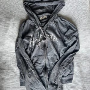 Grå hoodie från Hollister med dragkedja - Mysig grå hoodie från Hollister med broderad logga framtill. Tröjan har huva med snörning, dragkedja och två fickor. Storlek XS men passar även S eller M.