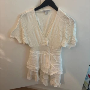 Vit kort omlottklänning från NewYorker - Superfin vit kortklänning från NewYorker med v-ringning, omlottkänsla och volangdetaljer. Klänningen har korta ärmar och är gjord i ett lätt, bomullstyg som känns perfekt för sommaren.