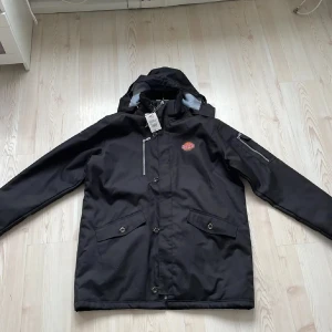 Svart vindjacka Printer Active Wear XL - Svart vindjacka från Printer Active Wear i storlek XL. Jackan har huva med grått foder, flera fickor med dragkedja och knappar samt en snygg logga på bröstet. Perfekt för kyliga dagar och har en sportig look med praktiska detaljer.