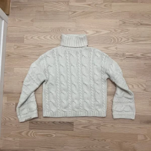 Vit stickad polotröja med flätmönster - Mysig vit stickad polotröja med grovt flätmönster och ribbade muddar. Tröjan har en hög krage och lång ärm, perfekt för kalla dagar. Den är lite boxig i modellen och passar dig som gillar en chill och avslappnad stil.