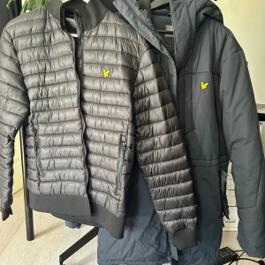 Grå/blå dunjacka och innerjacka från Lyle & Scott - Två  jackor från Lyle & Scott med gul logga på bröstet. Ena är en lång dunjacka med huva och dragkedjor på fickorna, perfekt för kalla dagar. Den andra är en kort pufferjacka med ribbade muddar och horisontella quiltade paneler, den går även att fästa på insidan av dunjackan.  Båda har dragkedja framtill. Båda jackorna för 800kr, 500kr var för sig.