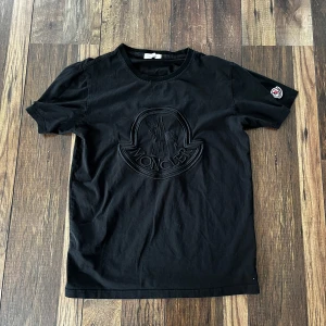 Svart Moncler t-shirt med broderad logga - Svart t-shirt från Moncler med stor, broderad logga ton-i-ton på bröstet och klassisk rund hals. Kortärmad modell i mjuk bomull med Moncler-märke på ena ärmen. Perfekt för dig som gillar stilrena och exklusiva plagg.