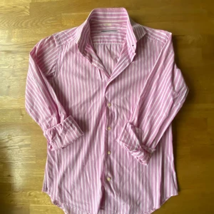 Rosa och vit randig skjorta Confezione Artigianale - Snygg skjorta i rosa och vitt med vertikala ränder från Confezione Artigianale. Klassisk krage, långärmad och knäppning framtill med ljusa knappar. Skjortan är tillverkad i mjuk bomull och har en normal passform som funkar till många olika stilar.