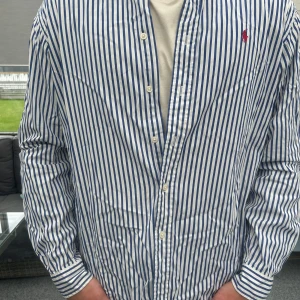 Ralph Lauren Classic Fit skjorta – blå/vit randig, stl L - Tidlös Ralph Lauren-skjorta i blå/vit rand med röd Polo-logo. Classic Fit stl L – rymlig och bekväm passform. 100% bomull, passar både vardag och fest. I fint skick och redo för ny ägare som gillar klassisk preppy stil. #ralphlauren #poloralphlauren #skjorta #herrskjorta #blåskjorta #randigskjorta #preppy #klassiskstil #secondhand #plickfashion #plickstyle #bomullsskjorta 