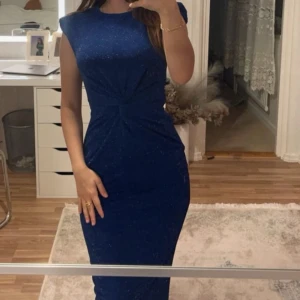 Marinblå glittrig långklänning - Snygg marinblå långklänning med glittrigt tyg och tight passform. Klänningen har en twistad detalj framtill i midjan och är ärmlös med rund halsringning. Perfekt för dig som vill sticka ut med en elegant och modern look.