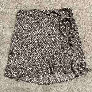 Brun blommig omlottkjol från Bikbok - Supersöt brun omlottkjol från Bikbok i storlek XS med små vita och gula blommor. Kjolen har volangkant nedtill och knytband i midjan. Material: viscose. Super fint skick! 