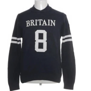 Mörkblå stickad tröja Morris Britain - Snygg mörkblå stickad tröja från Morris med vit text 'BRITAIN' och siffran 8 på bröstet. Två vita ränder på ärmarna ger sportig vibe. Union Jack-patch på ena ärmen. Rund hals och ribbade muddar. Perfekt för dig som gillar klassisk brittisk stil.