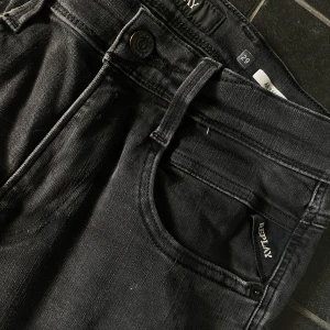 Svarta Replay jeansbyxor - Säljer ett par svarta jeans från Replay i modellen Anbass. Byxorna har klassisk femficksdesign, knapp och dragkedja framtill samt Replay-logga vid fickan. Passformen är slim och materialet är stretchigt jeans med lite slitningar på.
