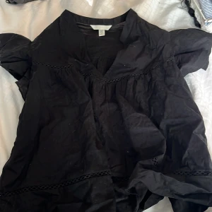 Svart blus från H&M - Svart blus  från H&M i storlek M. Modellen är kort med korta puffärmar och har dekorativa spetsdetaljer vid halsringningen och nedtill. Blusen har en loose passform och är tillverkad i bomull, vilket gör den skön och luftig.
