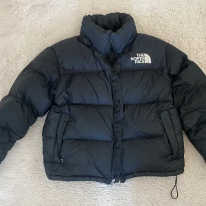 Svart pufferjacka från The North Face - Säljer en svart pufferjacka från The North Face, modell 700. Jackan har hög krage, två sidofickor med dragkedja och en snygg, boxig passform. Perfekt för kalla dagar med sin dunfyllda insida och klassiska logga på bröstet.