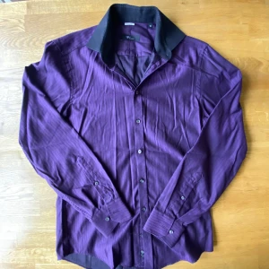 Lila randig skjorta från Venti - Snygg lila skjorta från Venti med diskreta ränder och svart kontrastkrage. Skjortan har lång ärm, klassisk krage och knappar hela vägen framtill. Perfekt för dig som vill sticka ut med färg och stil. Passar dig som gillar en lite mer dressad look.