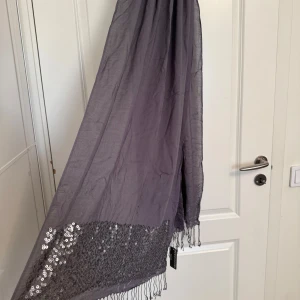 Grå glittrig halsduk från Kappahl - Snygg grå halsduk från Kappahl med glittriga paljetter längs ena kanten och tunna fransar i ändarna. Halsduken är rektangulär och har en lätt, luftig känsla som gör den enkel att styla till olika outfits. Perfekt för dig som gillar lite extra bling.