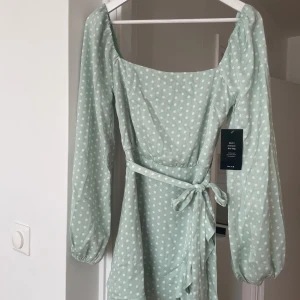 Mintgrön prickig klänning NA-KD - Superfin mintgrön kortklänning från NA-KD x Aïda Teller med vita prickar, långa puffärmar och knytband i midjan. Klänningen har omlott-detalj, dragkedja i sidan.
