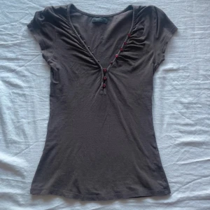 Brun v-ringad topp från Vero Moda - Brun topp från Vero Moda i storlek S med v-ringning och röda knappar längs halsen. 🩷