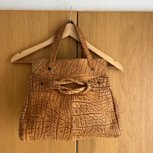 Brun handväska med mönster - Unik handväska i brun mocka med ett coolt, grafiskt mönster som påminner om krokodil. Väskan har två handtag, dekorativt bälte framtill och svarta nitar. Rymlig och perfekt för dig som gillar statement-accessoarer. I skinn.