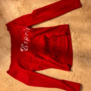 Röd långärmad topp från Esprit - Säljer en röd långärmad topp från Esprit med tryck och paljettdetaljer framtill. Tröjan har bred halsringning och coola grafiska texter i svart och silver. Perfekt för dig som gillar att sticka ut med färg och detaljer. Klippt till off shoulder 