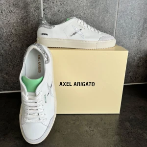 Axel Arigato vita sneakers med glitter - Snygga vita sneakers från Axel Arigato med silverglittrig häl och broderad fågel på sidan. Skorna har snörning, platt sula och detaljer i ljusgrönt på insidan. Inte alls använda , som man kan se på sulan. Pris kan diskuteras.