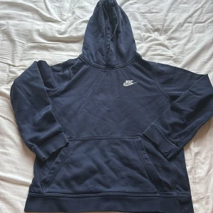Mörkblå Nike hoodie med huva - Snygg mörkblå hoodie från Nike med broderad vit logga på bröstet. Klassisk känguruficka framtill och skön huva. Tröjan är långärmad och har ribbade muddar vid ärmslut och nederkant. Perfekt för chill eller sportiga dagar.