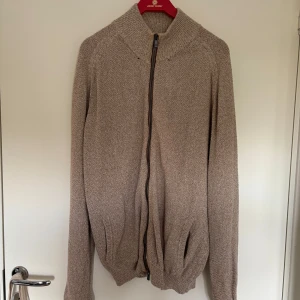 Beige kofta med dragkedja Massimo Dutti - Säljer en beige stickad kofta från Massimo Dutti med hög krage och hel dragkedja framtill. Materialet är en skön mix av bomull och linne, perfekt för lager-på-lager. Långärmad och med ribbade muddar för extra chill vibe. Snyggt basic-plagg som funkar till allt. Har pyttesmå hål, se bild 4 och 5 men bör vara lätta att laga!