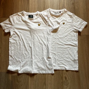 Vita Lyle & Scott t-shirts - Två vita t-shirts från Lyle & Scott i klassisk modell med rund hals och korta ärmar. På bröstet finns den ikoniska gula fågel-loggan som ger en clean och stilren vibe. Perfekt för dig som gillar enkel och snygg stil. Materialet är mjuk bomull.