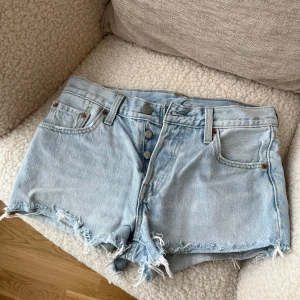 Levi's ljusblå jeansshorts med fransar - Snygga ljusblå jeansshorts från Levi's med klassisk femficksdesign och rå fransad kant. Shortsen har hög midja, knappgylf och cool vintage-vibe. Perfekta för varma dagar och festivalhäng. Levi's patch baktill och ikoniska röda etikett på fickan. Skulle beskriva storleken som XS/ XXS. Jag bär storlek S och de är cirka två storlekar för små för mig 