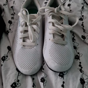 Puma AMG Petronas vita sneakers - Säljer ett par vita Puma AMG Petronas Formula One Team sneakers med svarta detaljer och turkosa loggor. Skorna har perforerat mönster, snörning och rund tå. Mercedes-emblem på hälen och platt sula. Perfekt för dig som gillar motorsport och stilren design.