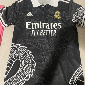 Real Madrid svart drakmönstrad tröja - Snygg svart Real Madrid fotbollströja från Adidas med coolt drakmönster i grått över hela tröjan. Vit krage och vita detaljer på ärmarna, samt klubbmärke och Adidas-logga på bröstet. Materialet är lätt och andas, perfekt för match eller träning.