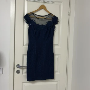 Marinblå glittrig kortklänning med mesh - Säljer en marinblå kortklänning med glittrigt tyg och mesh upptill. Klänningen har broderade detaljer med små pärlor och paljetter längs halsringningen. Modellen är ärmlös och har en slim passform som framhäver figuren. Perfekt för dig som vill sticka ut med snygga detaljer.